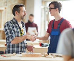 Foto: ein Ausbilder schüttelt einem Auszubildenden die Hand. Die beiden stehen an einer Werkbank in einer Holzwerkstatt. 