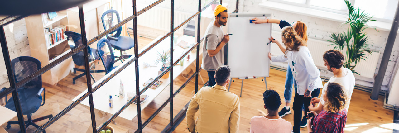 Foto: 7 Personen stehen im Kreis um eine Flipchart-Wand und erarbeiten gemeinsam Ideen.