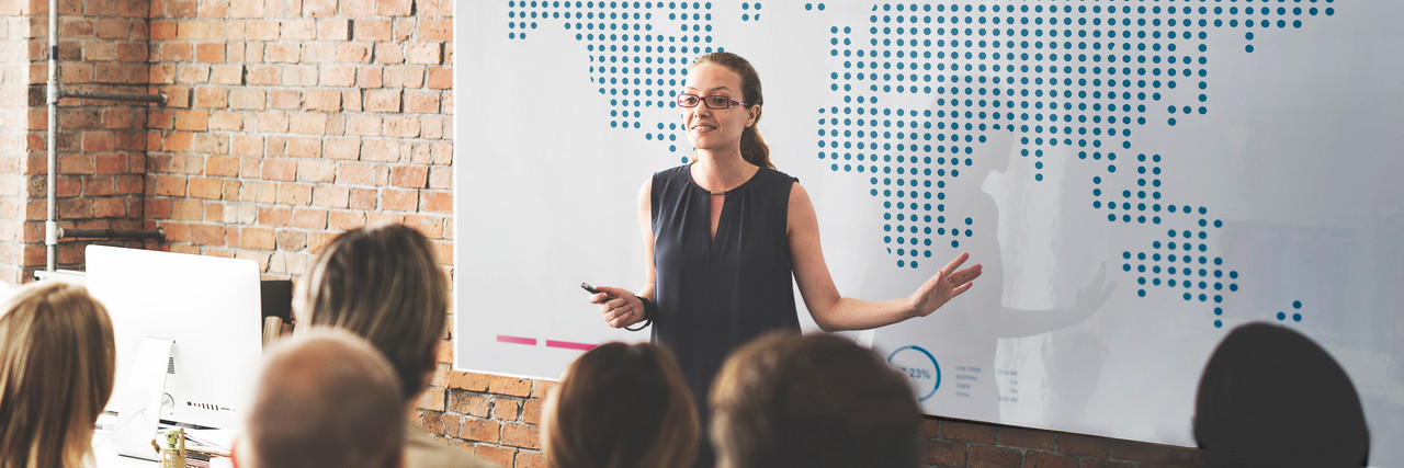 Foto: Eine Frau erklärt vor einer zuhörenden Gruppe am Whiteboard. Auf dem Whiteboard ist eine Weltkarte zu sehen.
