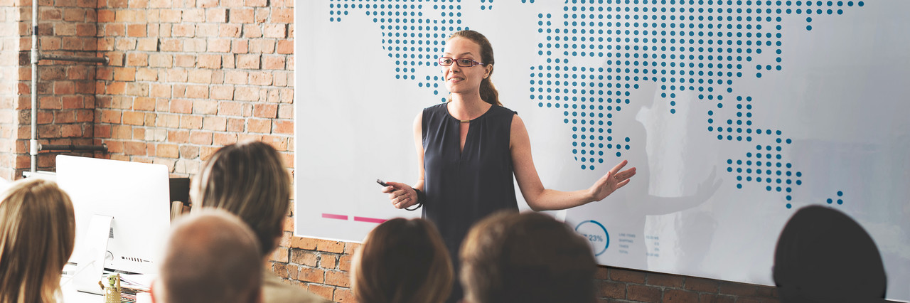 Foto: Eine Frau erkl&auml;rt vor einer zuh&ouml;renden Gruppe am Whiteboard. Auf dem Whiteboard ist eine Weltkarte zu sehen.