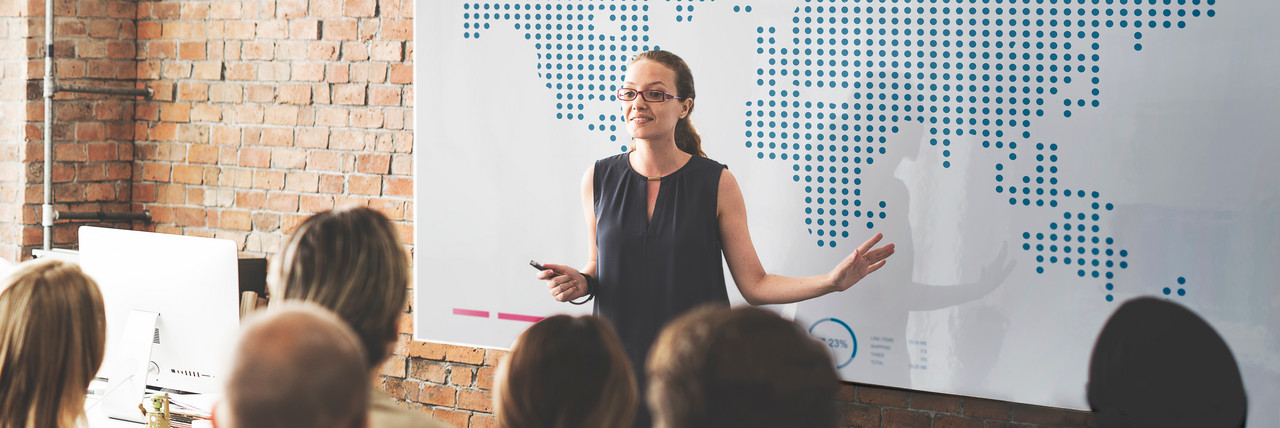 Foto: Eine Frau erklärt vor einer zuhörenden Gruppe am Whiteboard. Auf dem Whiteboard ist eine Weltkarte zu sehen.