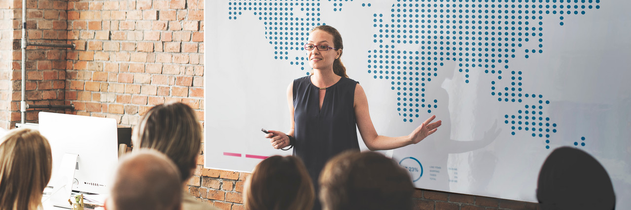 Foto: Eine Frau erkl&auml;rt vor einer zuh&ouml;renden Gruppe am Whiteboard. Auf dem Whiteboard ist eine Weltkarte zu sehen.