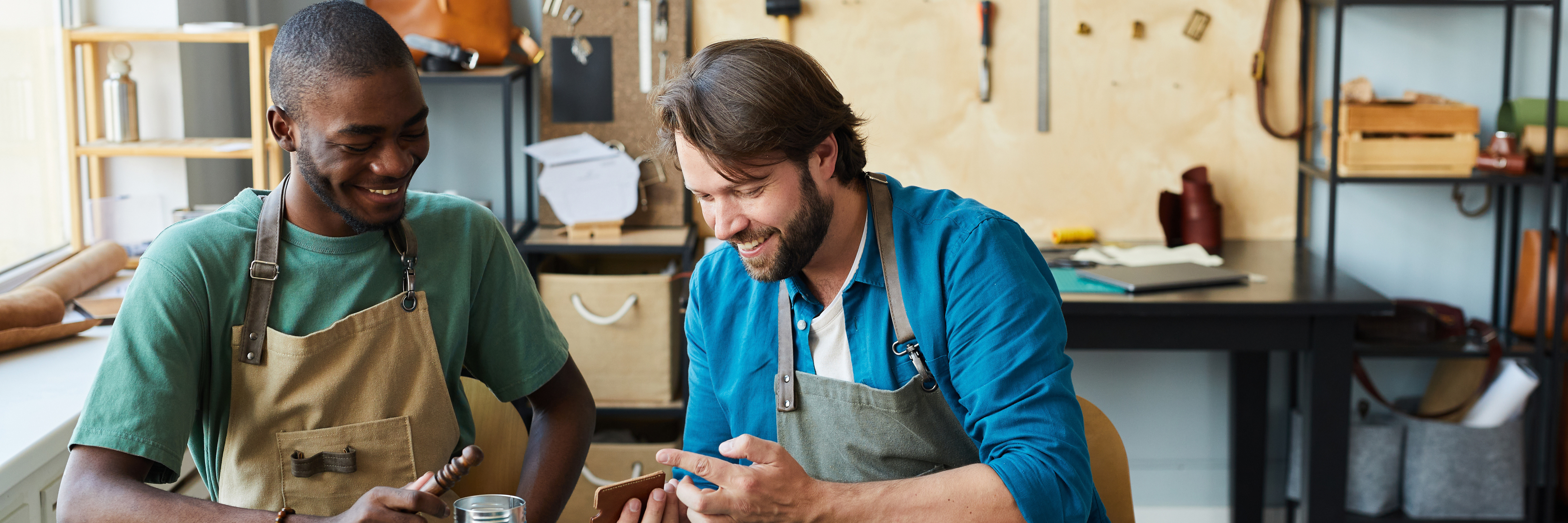 Foto: zwei junge Männer sitzen lächelnd und arbeitend an einer Werkbank und bearbeiten Leder.
