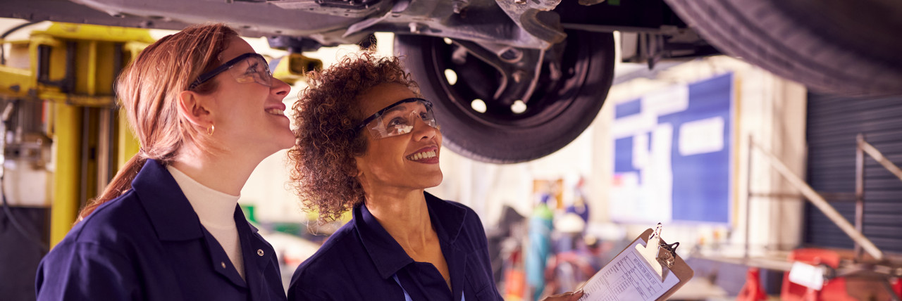 Foto: Ausbilderin erkl&auml;rt einer Auszubildenden den Autounterboden unter einem Auto, das auf der Hebeb&uuml;hne steht, stehend..