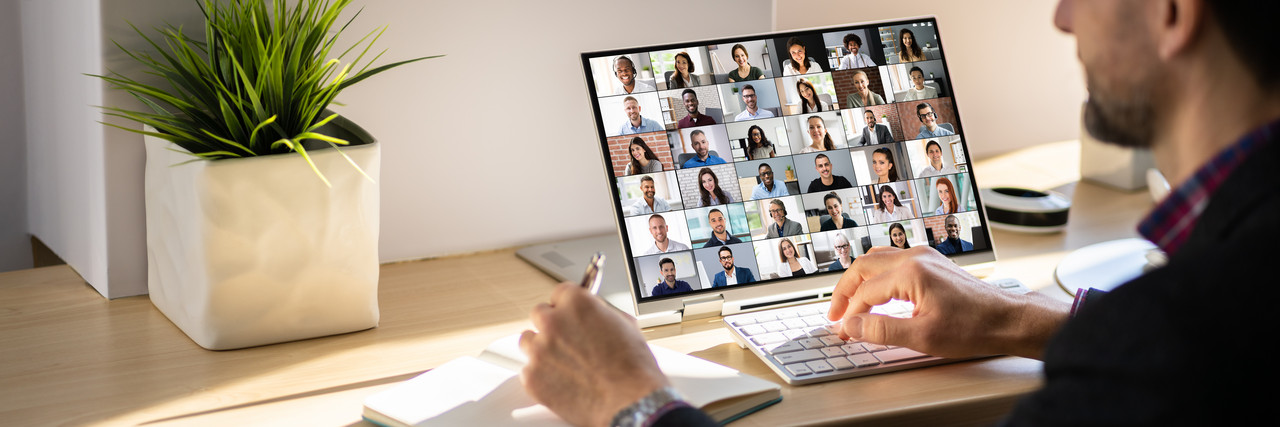 Foto: ein Mann sitzt vor dem Notebook. auf dem Bildschirm ist eine Videokonferenz mit vielen Videokacheln zu sehen. 