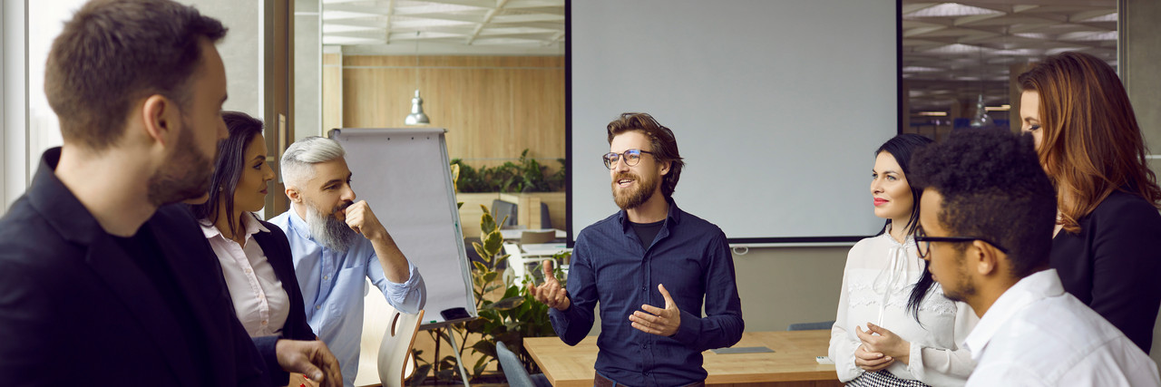 Foto: Menschen stehen in einem Büro vor einem leeren FlipChart und gestikulieren