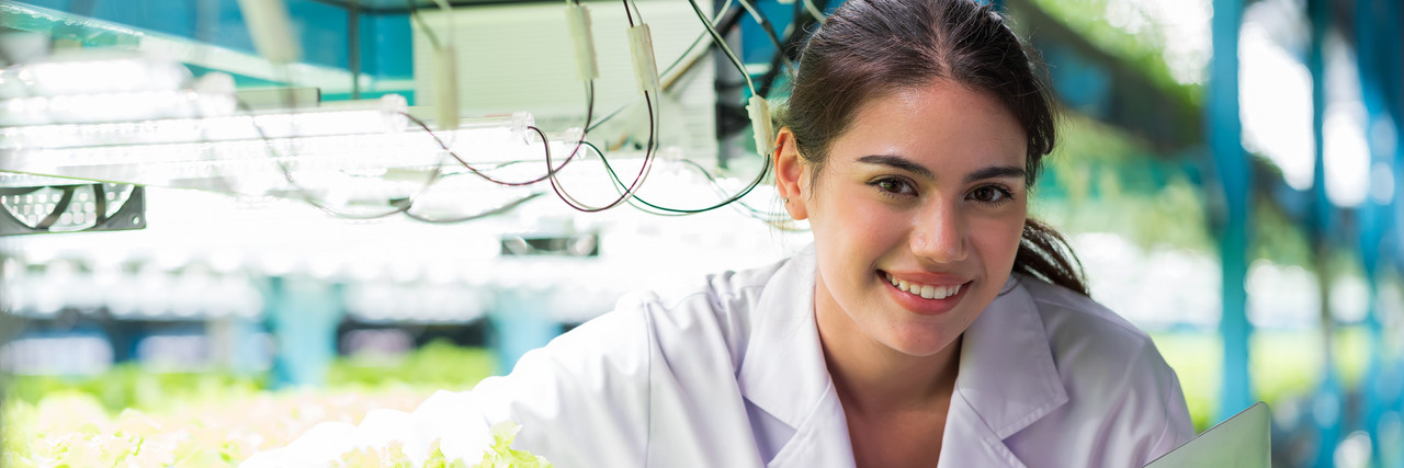 Foto: eine junge Auszubildende in Forschungsjacke neben einer Gem&uuml;seanbaufl&auml;che, die auf einer vertikalen Indoor-Farm angebaut werden.