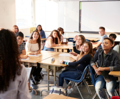 Foto: Eine gemischte Schulklasse, an Gruppentischen sitzend, schaut zur Lehrerin, deren Silhouette am linken Bildrand erkennbar ist.