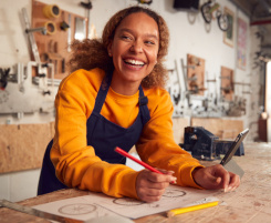 Foto: junge Frau in Werkstatt mit Stift und Papier, lacht in die Kamera