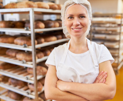 Foto: eine junge Ausbilderin im Backhandwerk lächelt vor einem rollbaren Regal mit gebackenen Broten in die Kamera.