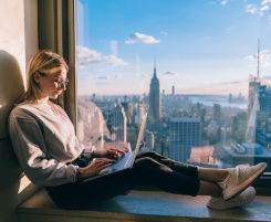 Foto: eine Auszubildende sitzt mit Notebook auf den Beinen auf der Fensterbank. Im Hintergrund ist die Skyline der Stadt New York zu sehen.