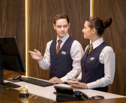 Foto: ein Hotelfachmann und eine Hotelfachfrau an der Hotelrezeption. Er erklärt ihr gerade etwas und zeigt auf den Bildschirm. 