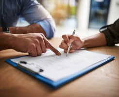 Foto: Im Mittelpunkt ein Klemmbrett mit Dokument. Daneben sitzen zwei Personen, deren Arme erkennbar sind. Die eine Hand hält einen Stift, die Hand der anderen Person zeigt auf das Blatt im Klemmbrett.