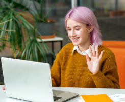 Foto: eine junge Auszubildende sitzt vor einem Notebook und winkt lächelnd in die Kamera.