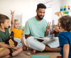 Foto: ein junger Erzieher in Ausbildung sitzt mit den Kindern in einem Kreis und liest aus einem Buch vor.