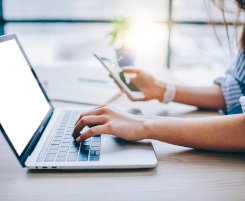 Foto: zwei Hande am Notebook. Eine Hand tippt auf dem Keyboard, in der anderen Hand ein Smartphone.